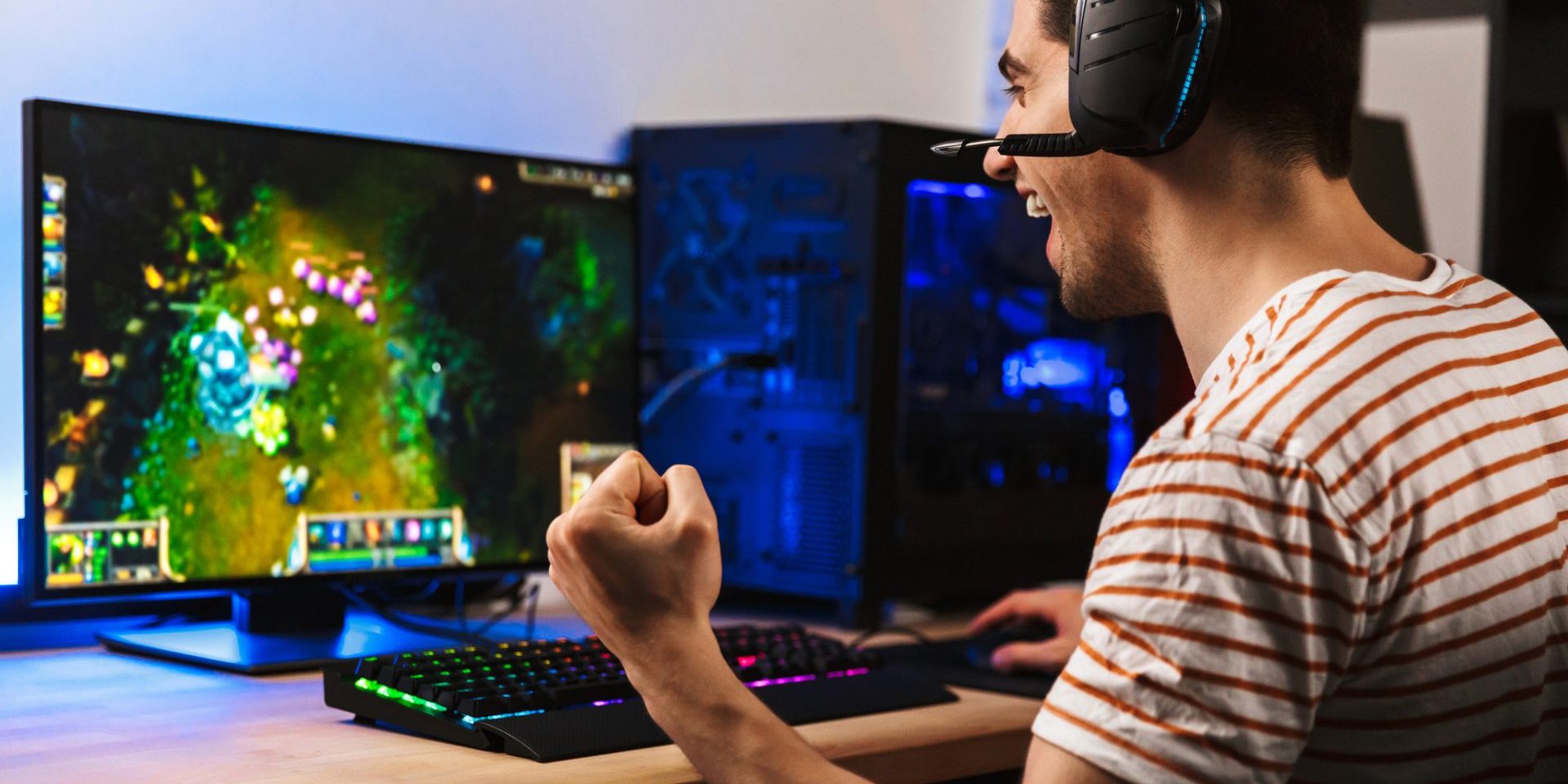 Portrait of delighted young guy playing video games on computer wearing headphones and using backlit colorful keyboard