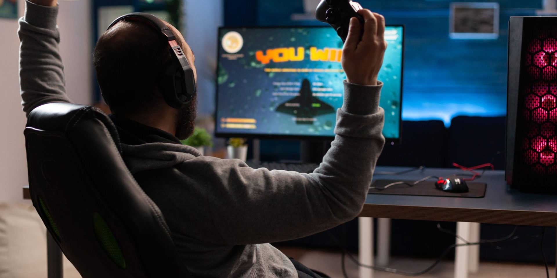 Man winning video games with joystick and headphones in front of monitor. Gamer using controller and headset, playing online games on computer. Person celebrating game win for leisure.