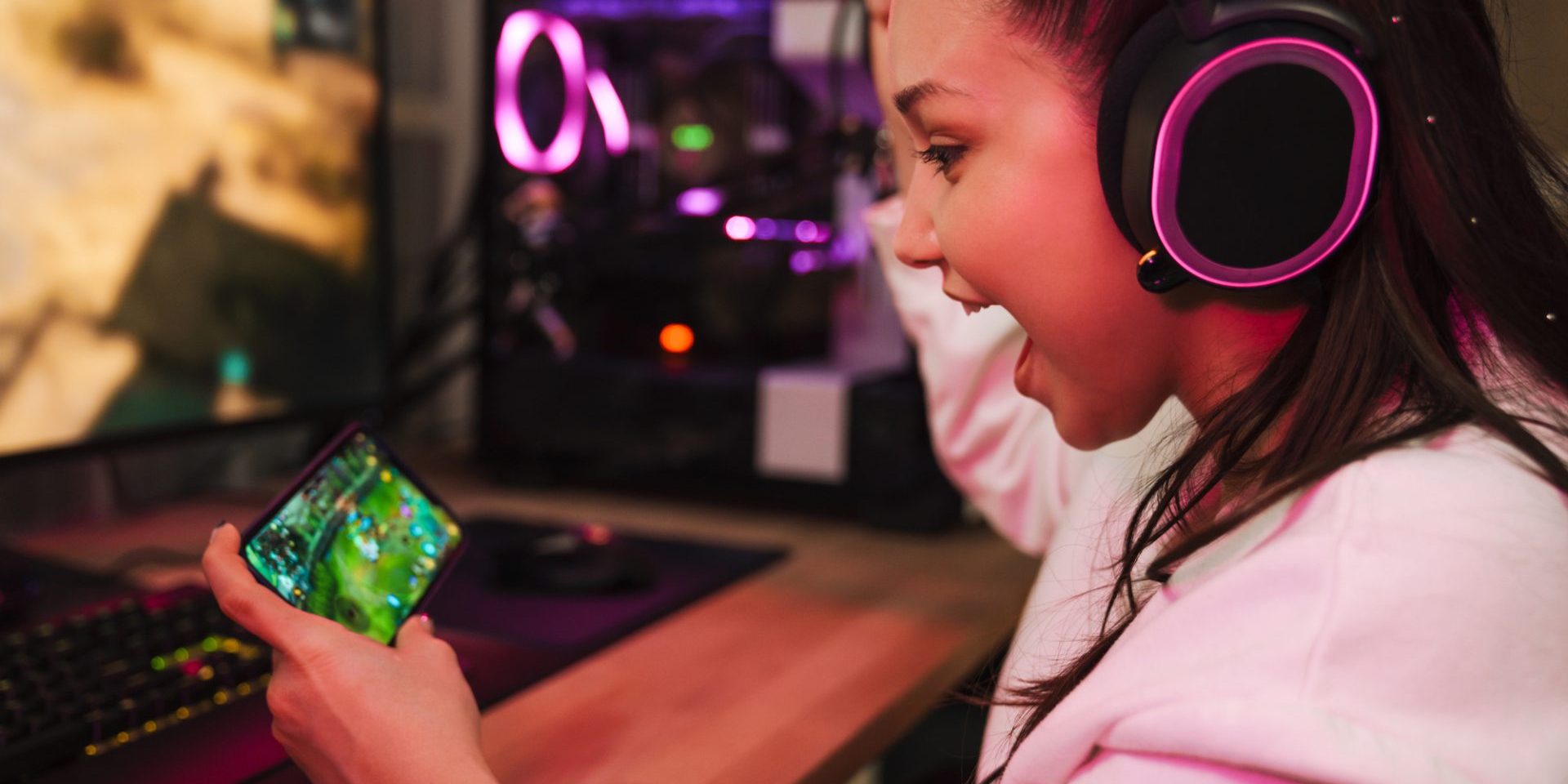 Young happy woman gamer playing video games on smartphone and computer in dark room wearing headphones