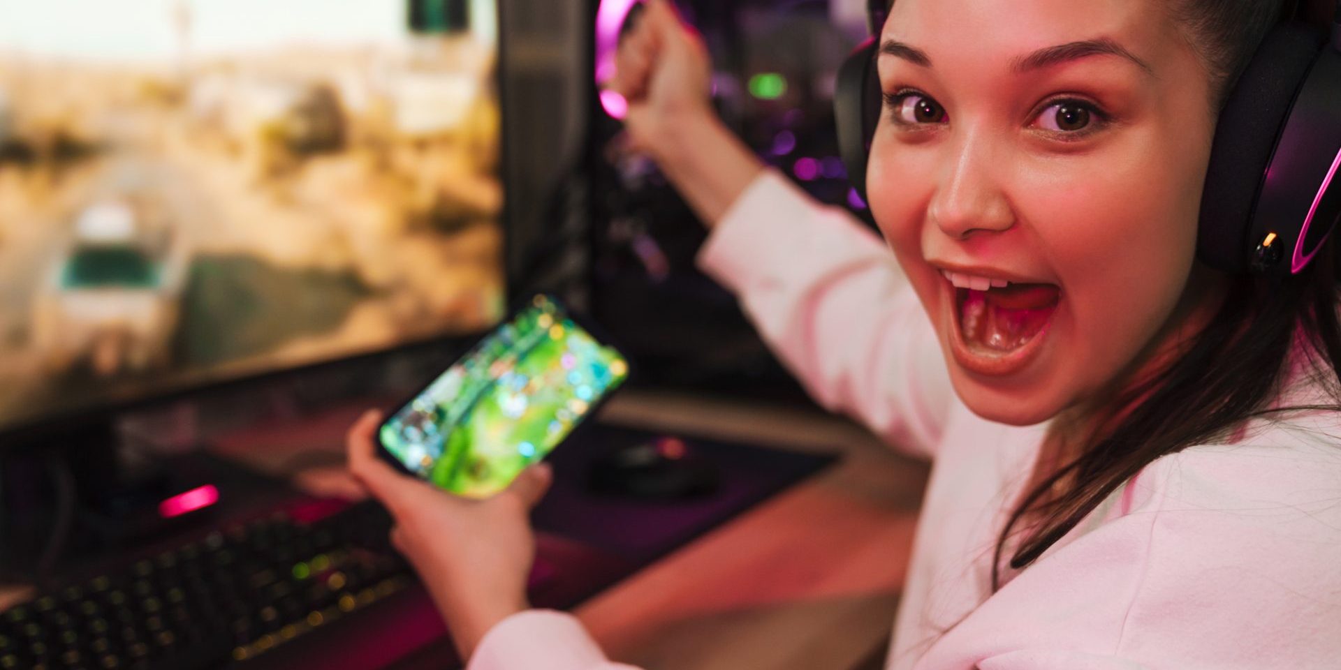 Young happy woman gamer playing video games on smartphone and computer in dark room wearing headphones