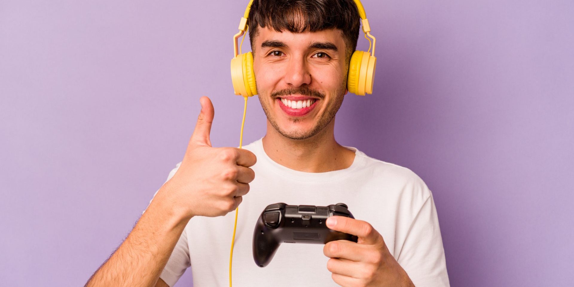 Young hispanic man playing with a video game controller isolated on purple background smiling and raising thumb up