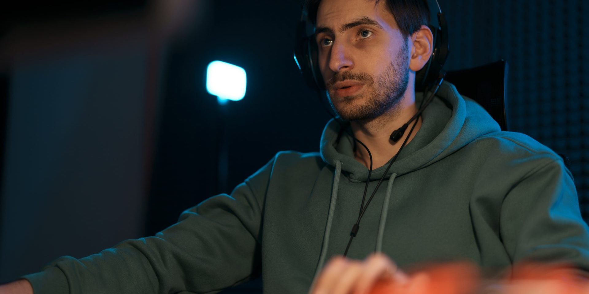 Concentrated male cybersport gamer wearing headset and playing video game having battle in dark room illuminated by neon lights