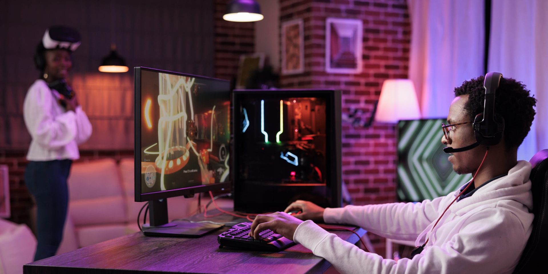 Man immersed in gaming session at computer desk while girlfriend in background uses virtual reality goggles. African american couple playing games, having fun on gaming pc and vr system